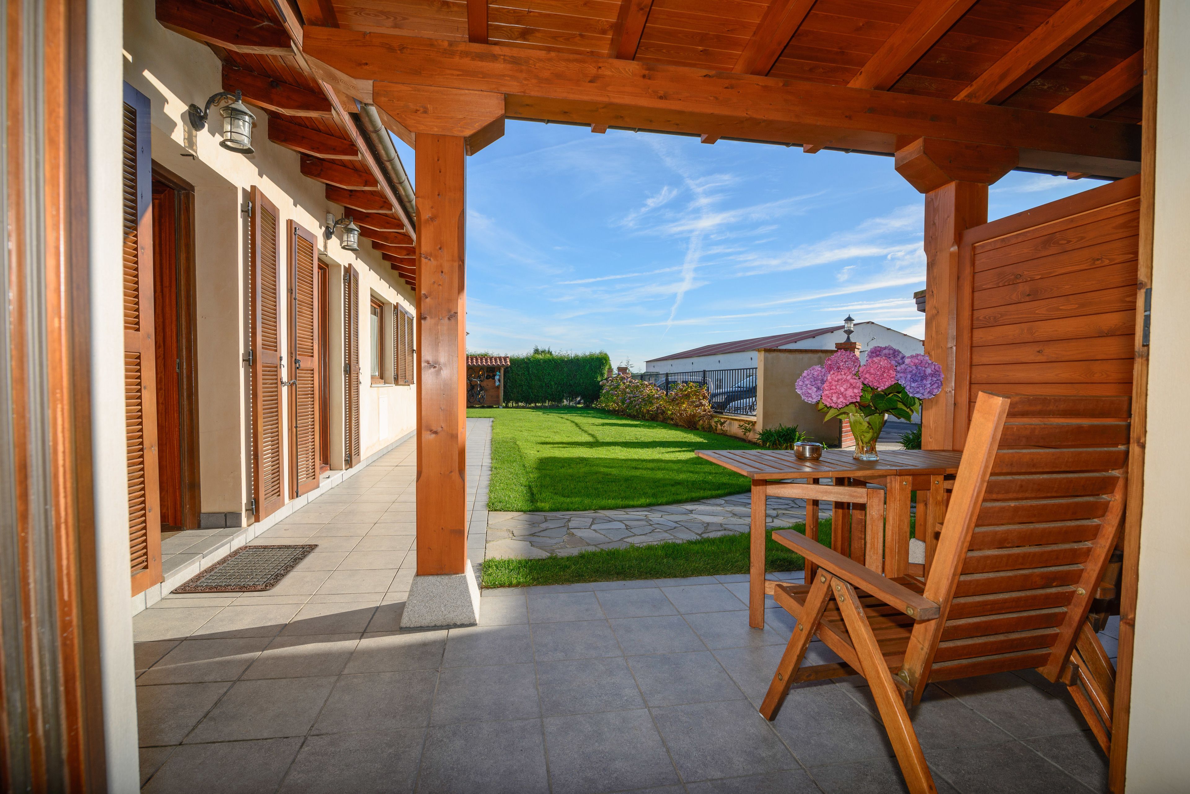 Een overdekt terras met een houten tafel en stoel, een kleine tuin met gazon en bloemen, en een gebouw met houten luiken op de achtergrond.