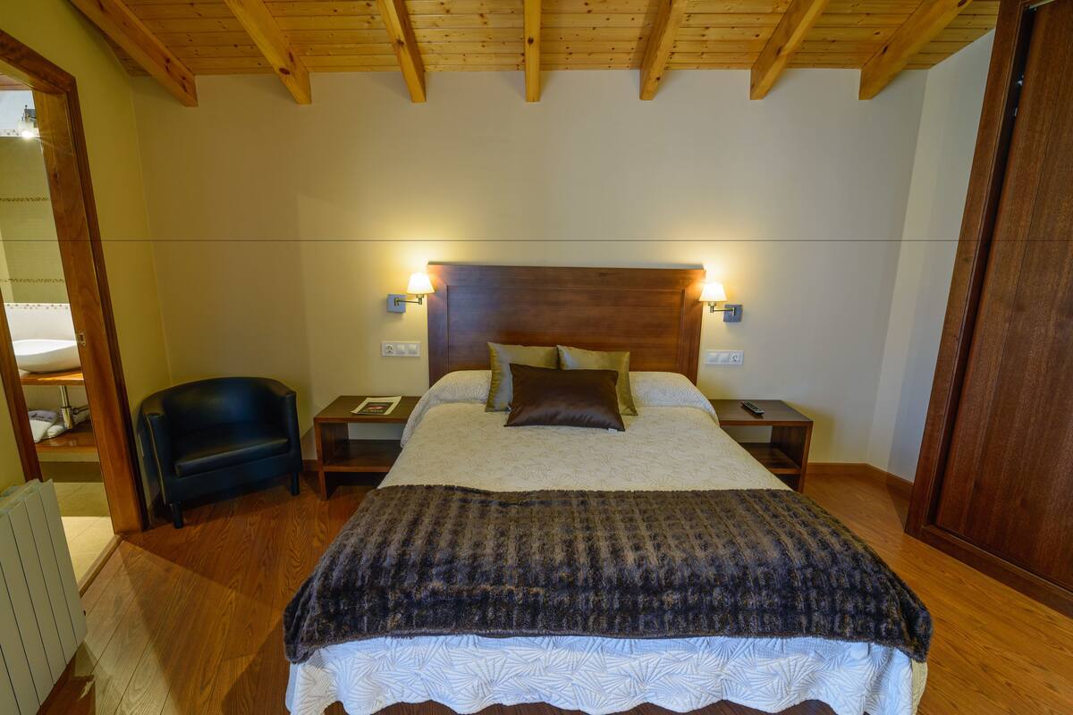 Une chambre avec un plafond en bois, un lit recouvert d’une couverture sombre, deux lampes de chevet, une table de chevet et un fauteuil en cuir noir.