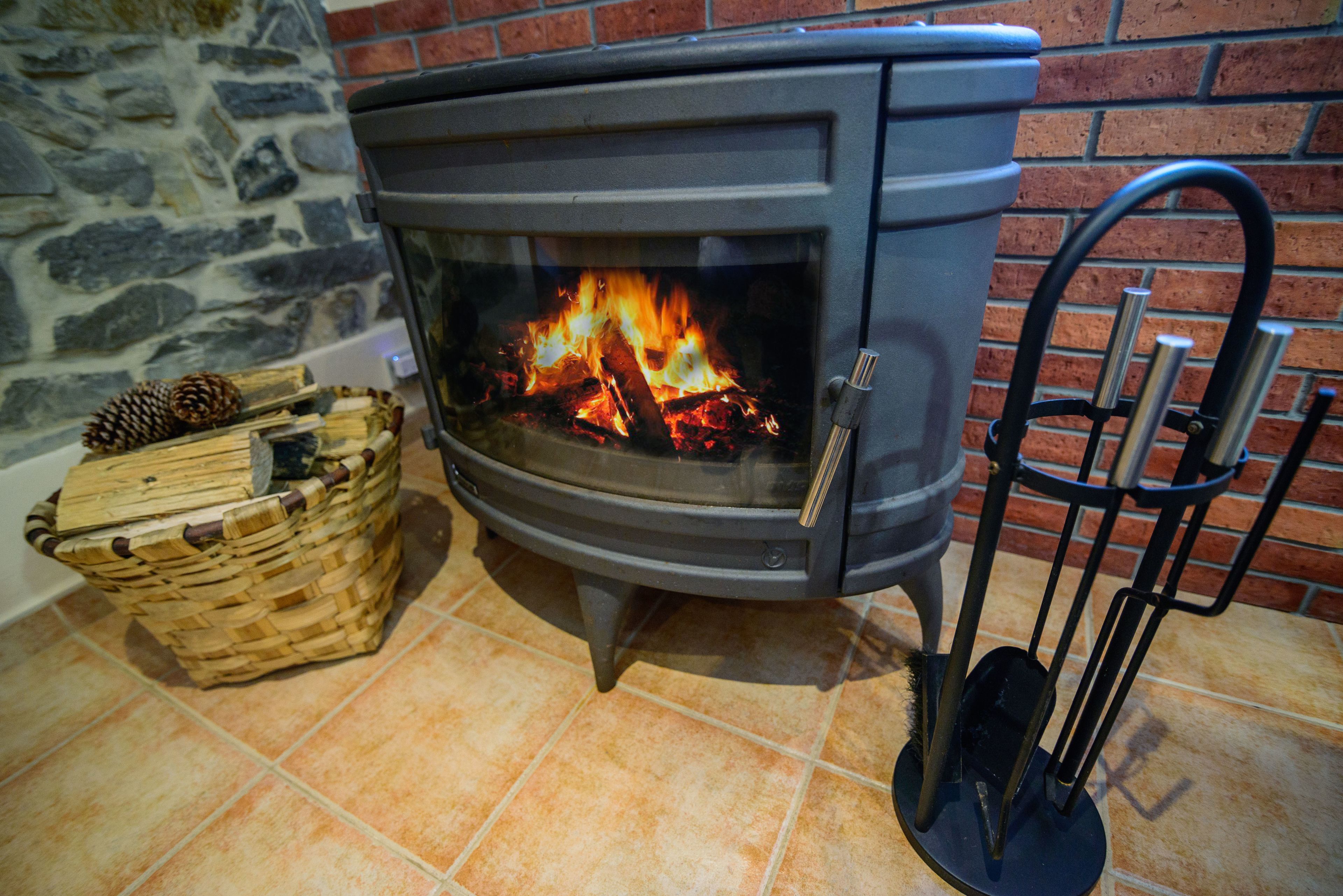 A wood-burning stove with a glass door, a basket with pinecones, and a metal ash collector.