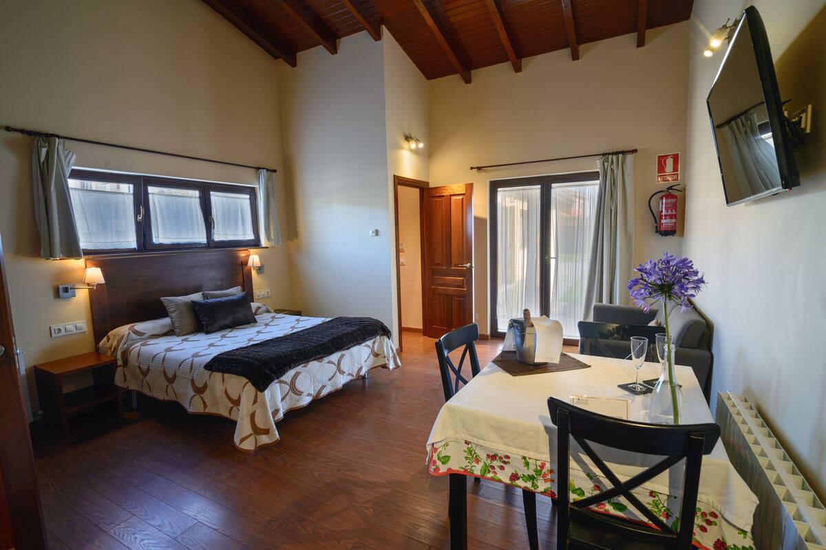 Une chambre à coucher comprenant un lit, des lampes de chevet, une tête de lit en bois, une fenêtre avec des rideaux, une porte et un coin repas avec une table et des chaises.