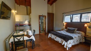 Un dormitorio con cama, mesita de noche y ventana. Se ve un área de cocina con armarios y encimera a través de una puerta abierta.