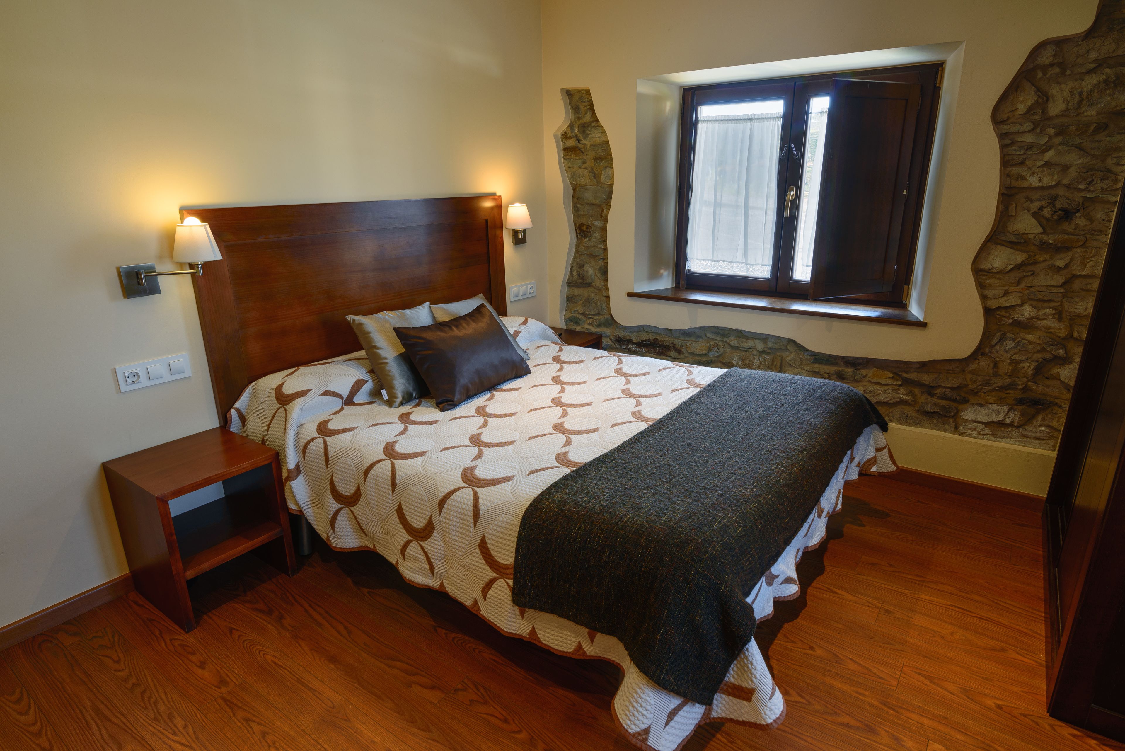 Un dormitorio con cabecera de madera, cama con ropa de cama estampada, mesita de noche, pared de acento de piedra y ventana con cortinas.