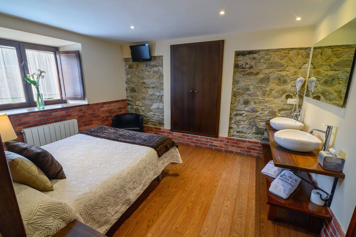 Une chambre à coucher avec un lit, un plancher en bois, un mur en pierre, une porte en bois, une salle de bain avec un lavabo et un miroir, et une fenêtre avec des rideaux.