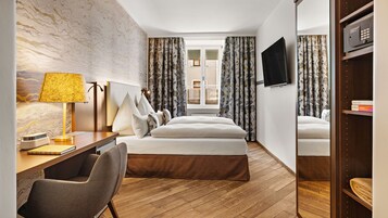 Ein Hotelzimmer mit einem Bett, einem Schreibtisch mit Lampe, einem Sessel, einem an der Wand befestigten Fernseher und Vorhängen mit Blättermuster.