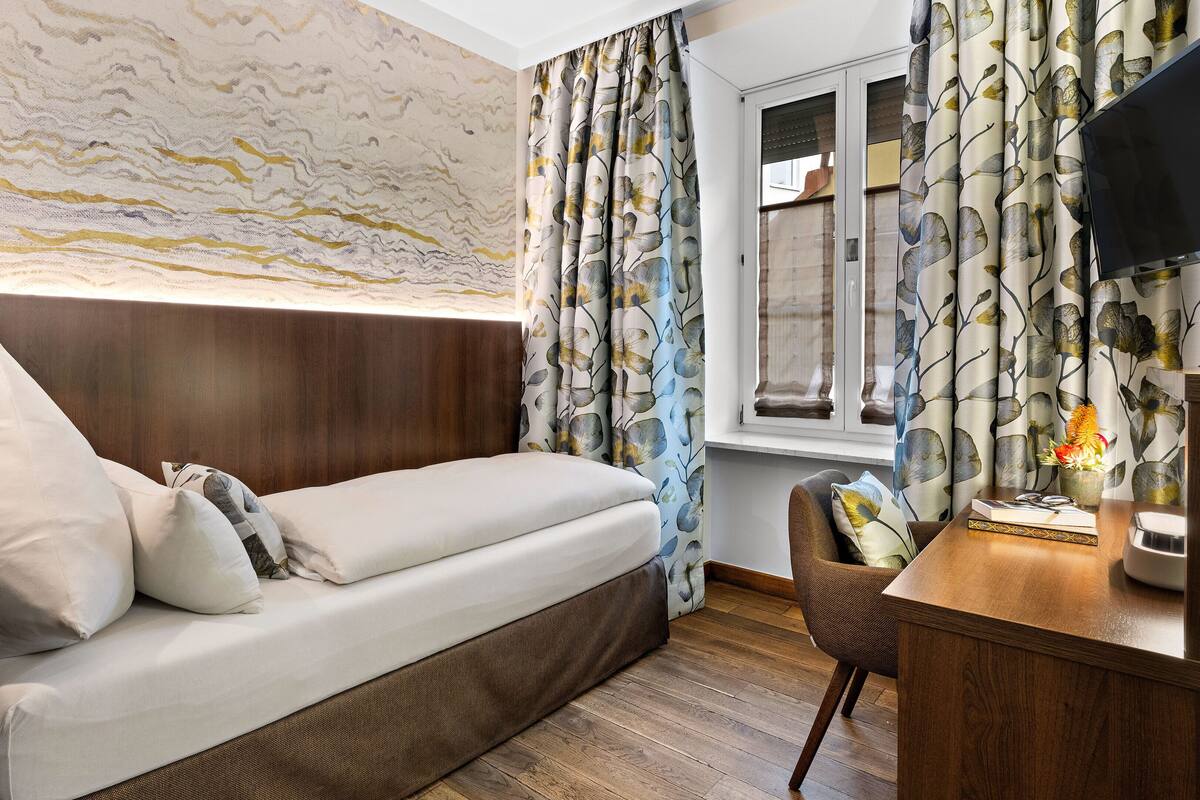 A hotel room with a bed, a desk, a chair, a window with curtains, and a wall with a marble-like pattern.