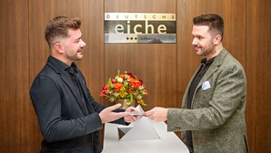 Two men exchanging a bouquet of flowers in front of a wooden wall with a sign that reads "DEUTSCHE eiche - SUPERIOR". - Hotel Deutsche Eiche (Munich)