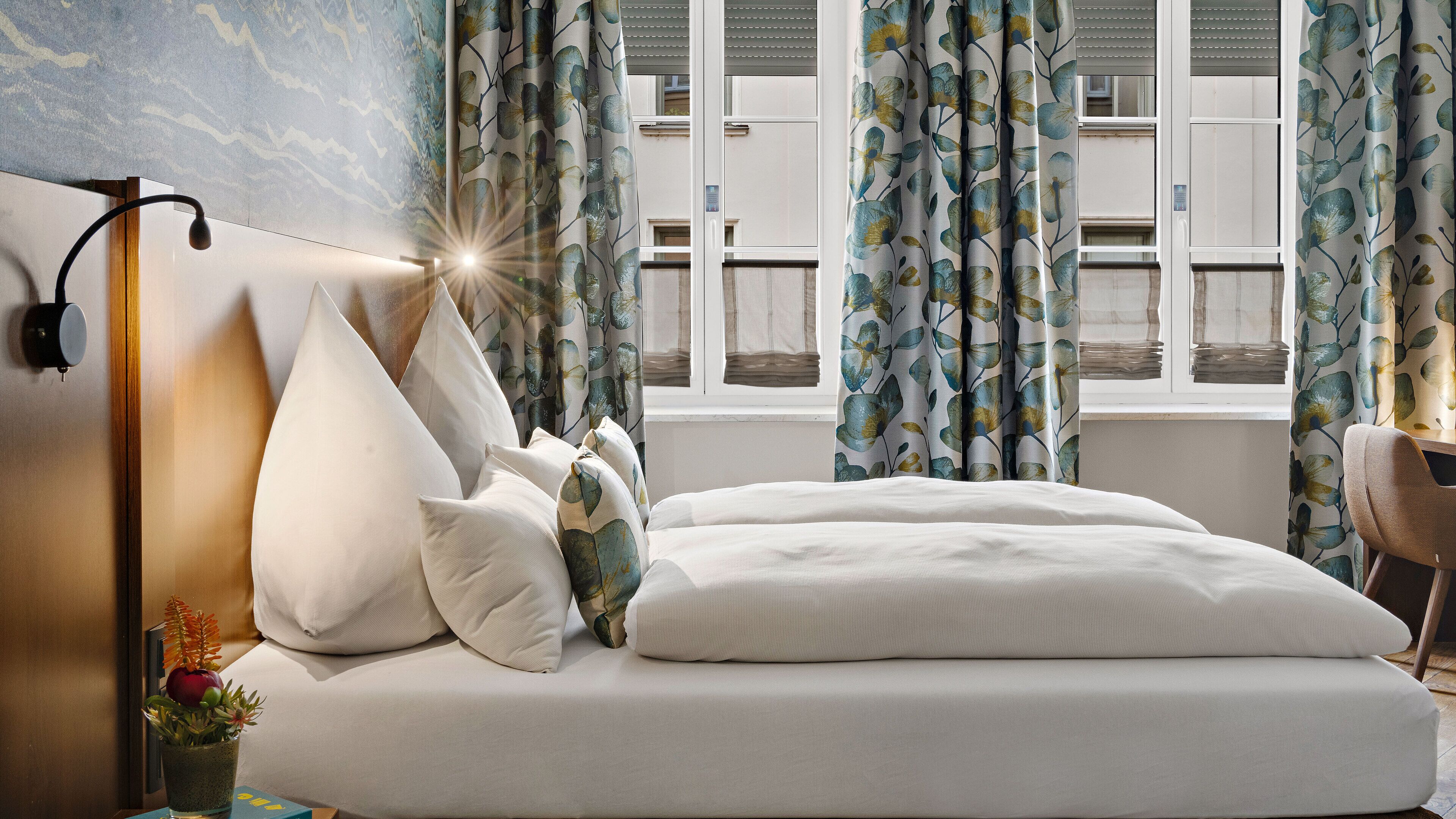 Una cama bien tendida con ropa de cama blanca y varias almohadas, una lámpara de noche y una pequeña planta en la mesita.
