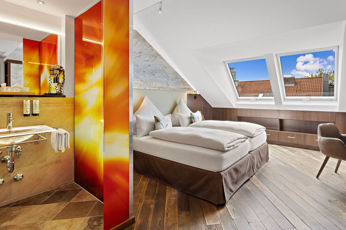 A modern bedroom with a skylight, a wooden floor, a bed, a desk, and a bathroom with a mirror and sink.