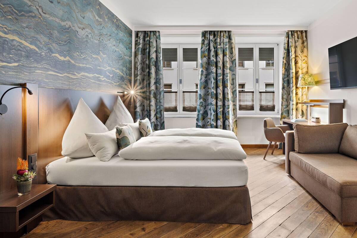 A modern hotel room with a large bed, a sofa, a desk, and a TV.