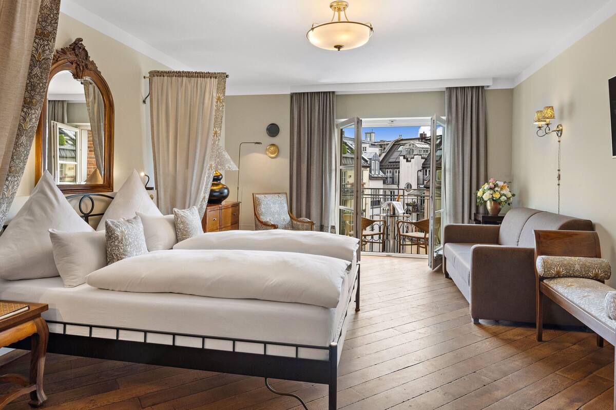 A bedroom with a bed, two chairs, a wooden table, a mirror, and a balcony with a view of buildings.