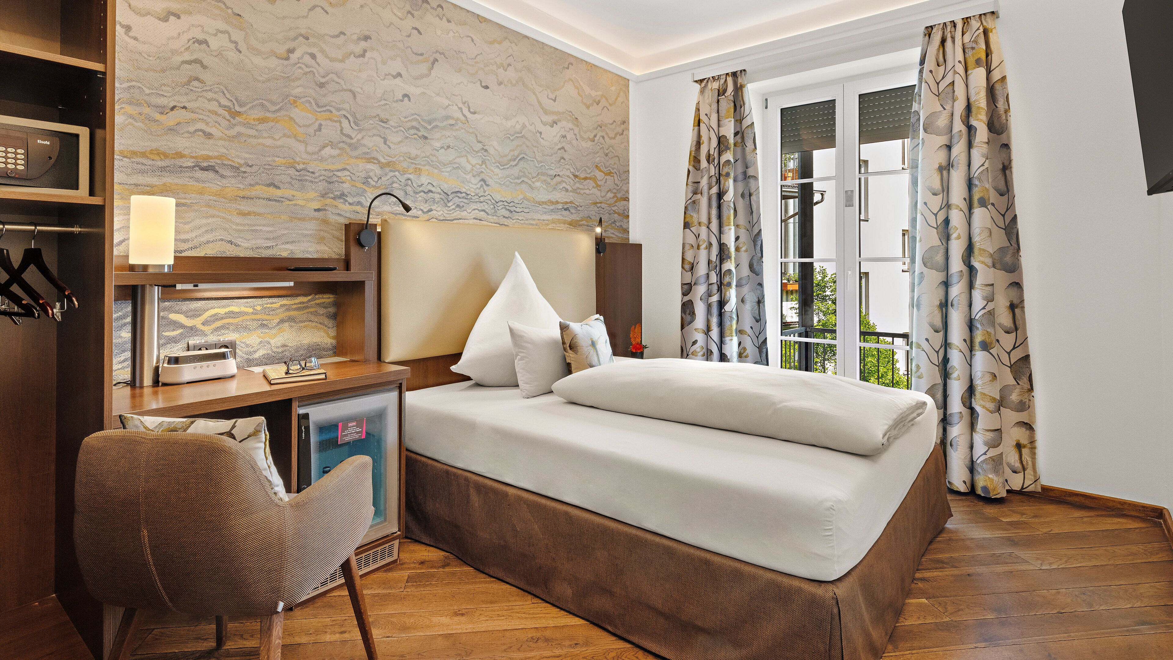 Un dormitorio con cama, mesita de noche, lámpara, silla y vistas al exterior.