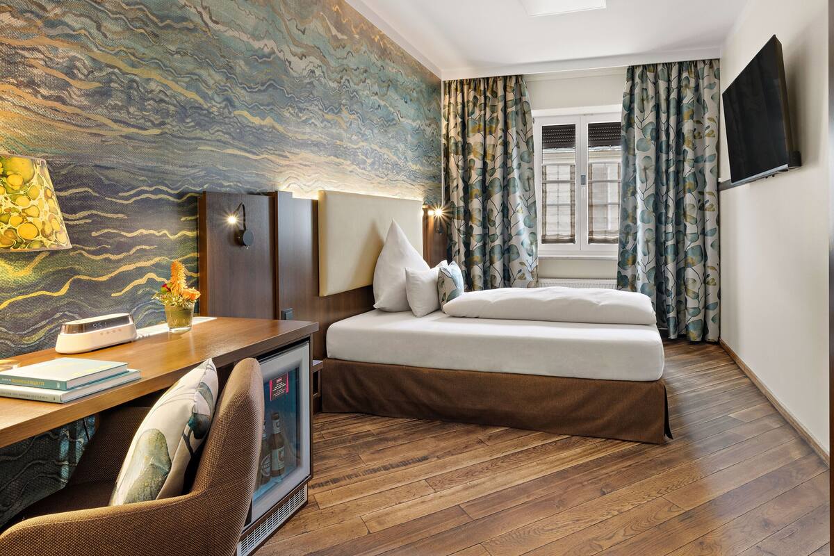 A modern hotel room with a large bed, a desk with a lamp, a television, and a large window with curtains.