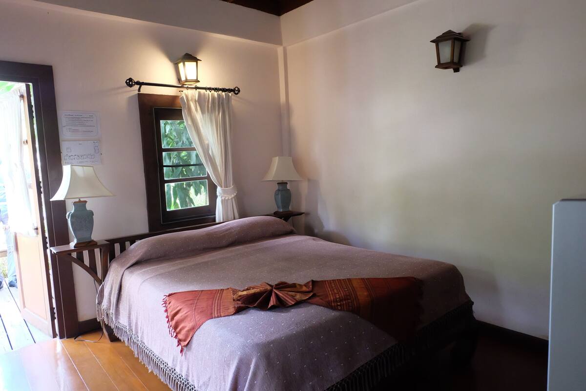 A neatly made bed with a patterned bedspread, two bedside lamps, a window with curtains, and a small wall-mounted lantern.