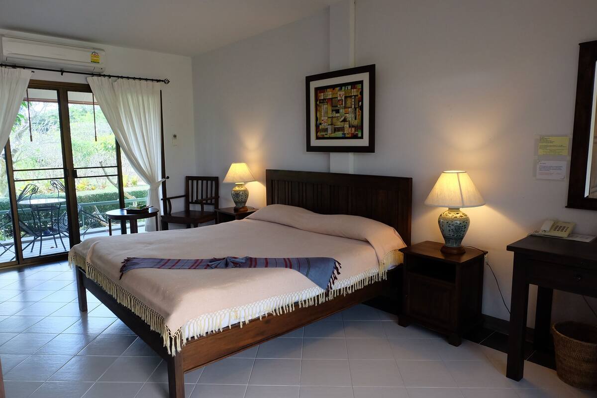 A bedroom with a bed, two bedside tables with lamps, a chair, a small table, a framed picture on the wall, and a balcony with a table and chairs.