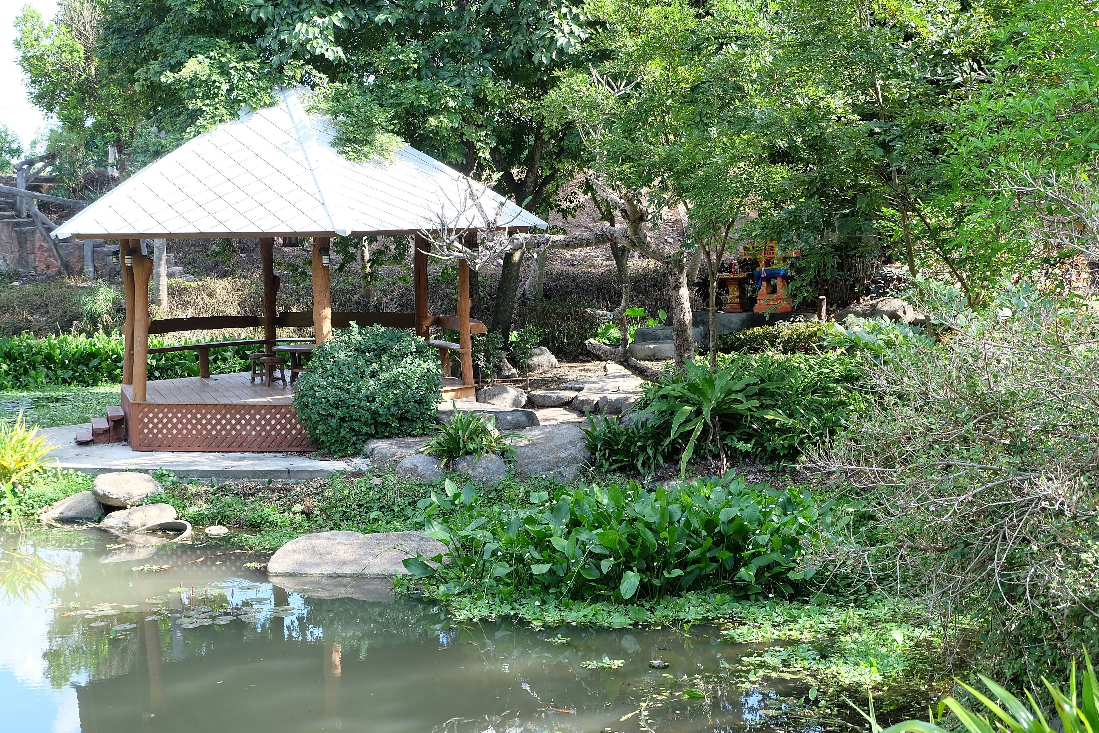 A gazebo with a tiled roof, surrounded by a pond, rocks, and lush greenery.