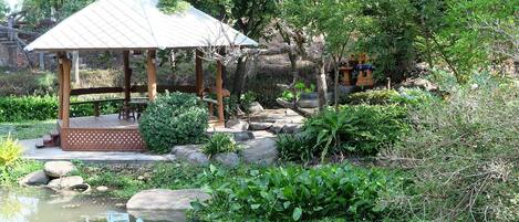 A gazebo with a tiled roof, surrounded by a pond, rocks, and lush greenery.
