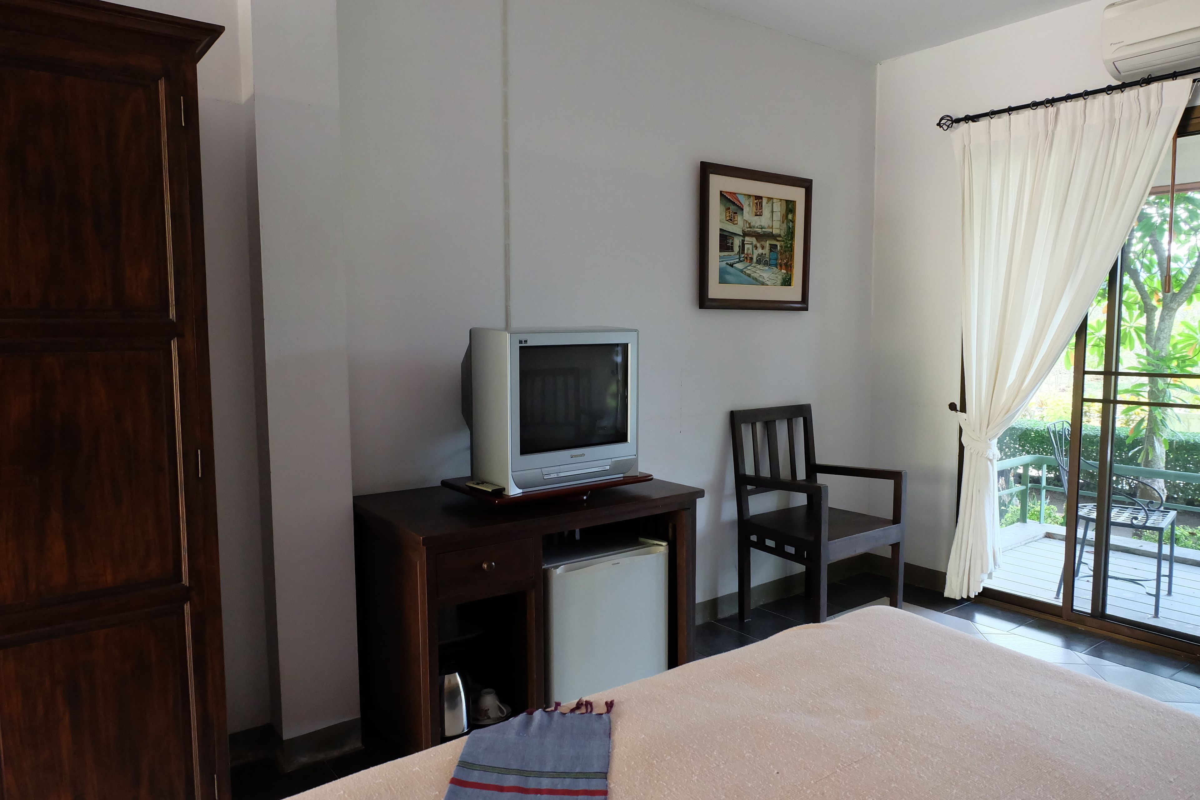 Una habitación de hotel con cama, televisor sobre un soporte de madera, una silla y una ventana con cortinas.