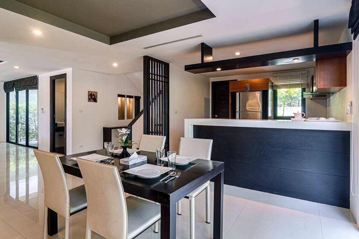 Une cuisine moderne avec un plan de travail noir et des appareils en acier inoxydable. On y trouve un coin repas avec une table noire et des chaises blanches.