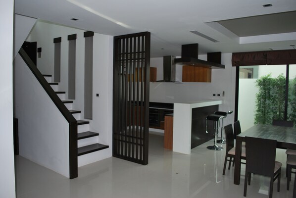 A modern kitchen with a dining area, a staircase, and a view of greenery outside. - Ocean Breeze Residence (Takua Pa)