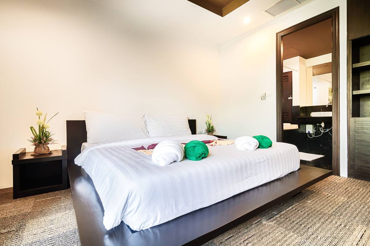 Une chambre d’hôtel moderne avec un grand lit, une table de chevet agrémentée d’une plante et un miroir dans la pièce adjacente.
