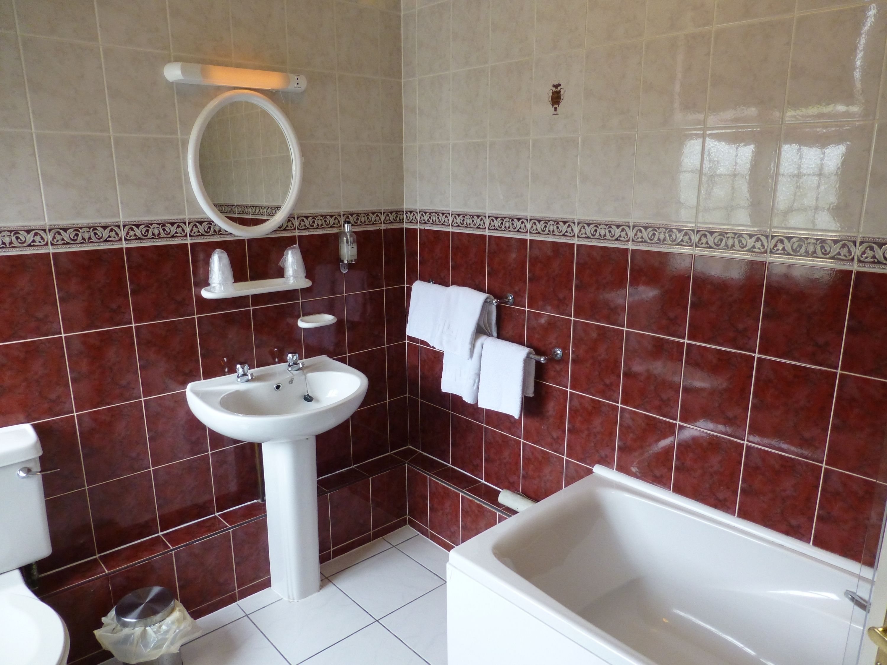 A bathroom with a white sink, a toilet, and a bathtub. There is a mirror above the sink and a towel rack with a towel hanging on it. The walls are tiled with a red pattern.