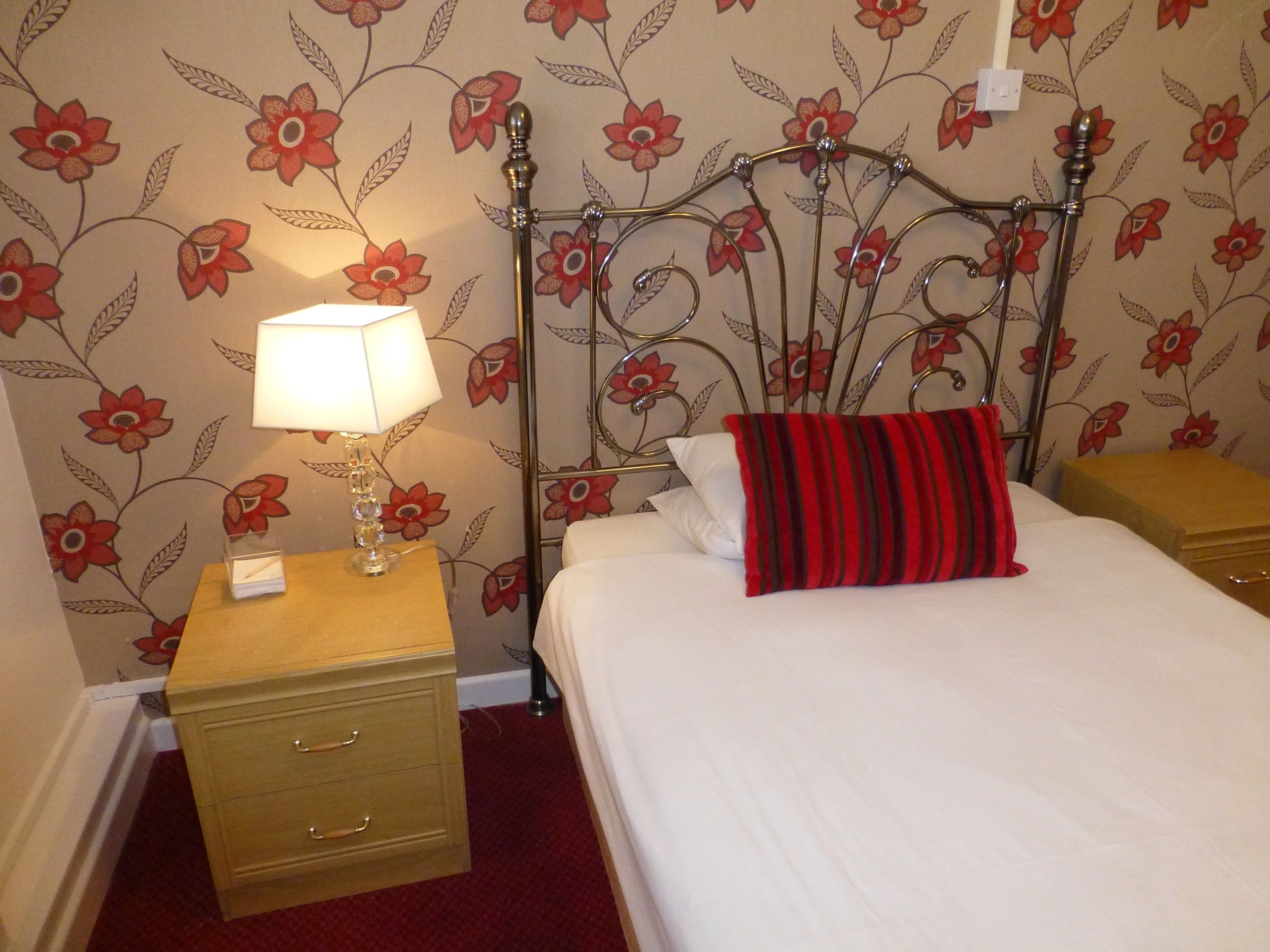 Una habitación de hotel con papel tapiz floral, estructura de cama metálica, mesita de noche con lámpara y almohada a rayas rojas y negras.