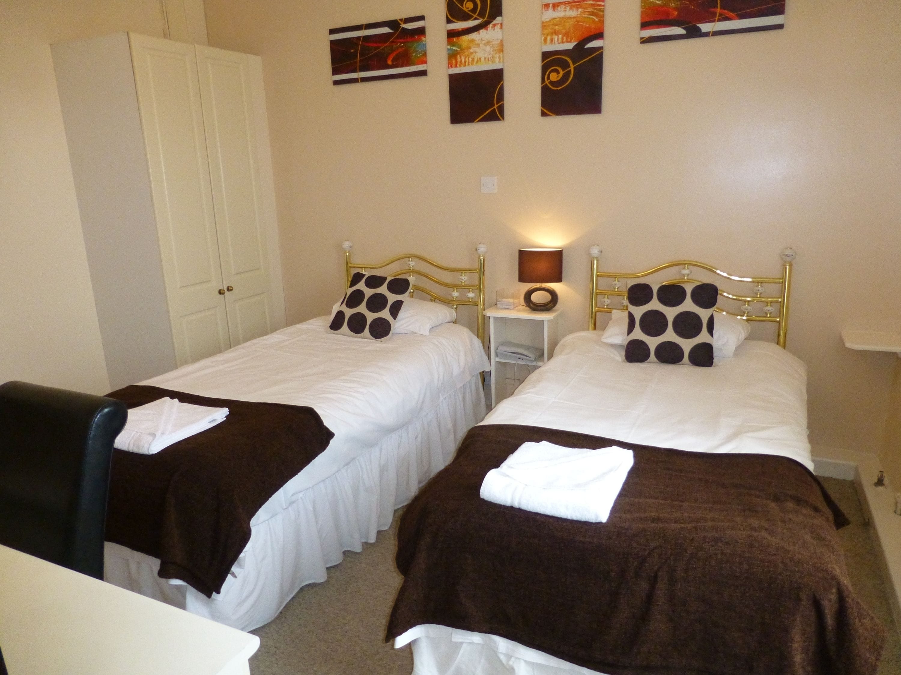A hotel room with two single beds, a wardrobe, a bedside table with a lamp, and artwork on the wall.