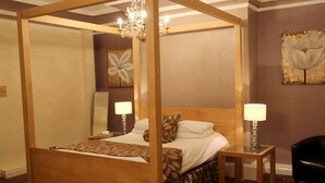 A four-poster bed with a wooden frame, two bedside tables with lamps, a large floral painting on the wall, and a black leather chair. - Maes Manor Hotel (Blackwood)