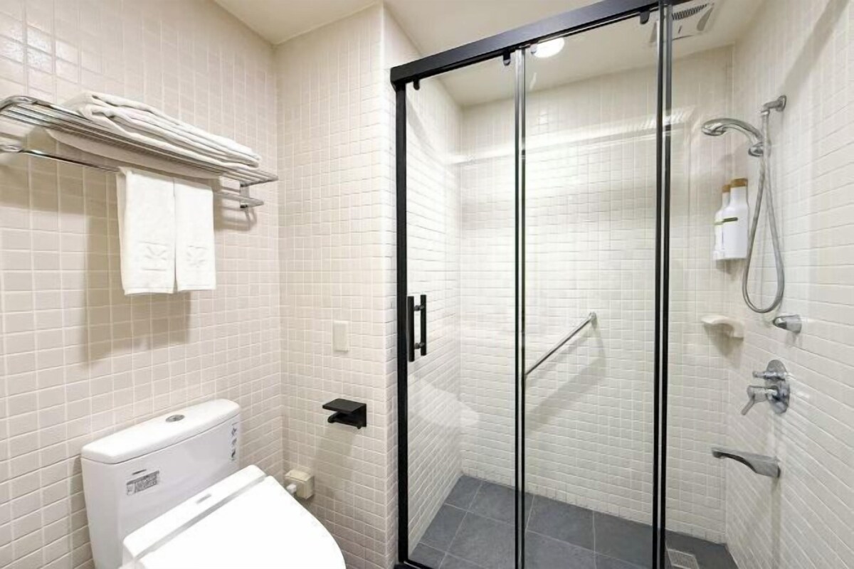 Une salle de bain moderne avec des murs recouverts de carreaux blancs, une douche à parois de verre, des toilettes blanches et un porte-serviettes.