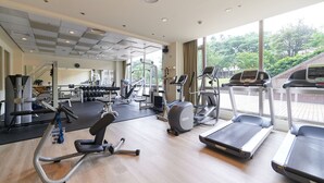 A gym with various exercise equipment, large windows, and a view of trees and a building outside. - Aspire Resort (Taoyuan City)