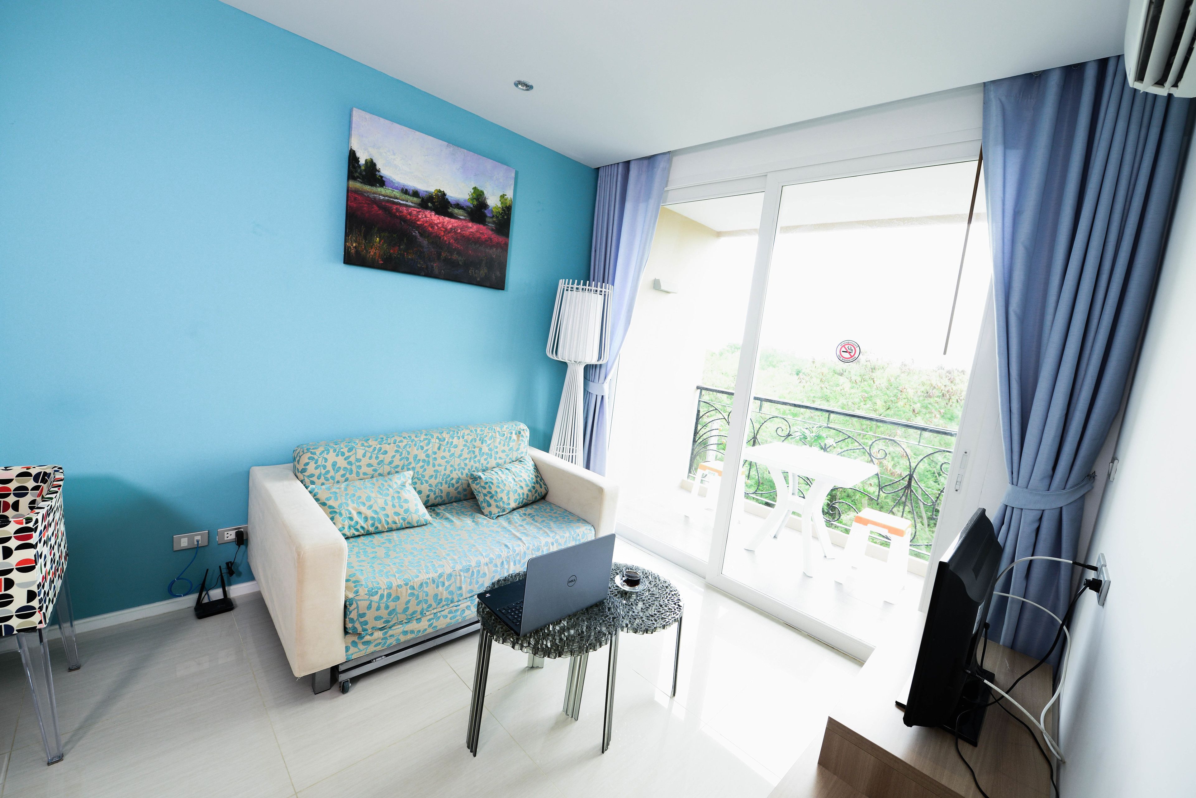 A modern living room with a blue wall, a white sofa with patterned cushions, a small round table with a laptop, and a balcony with a view.
