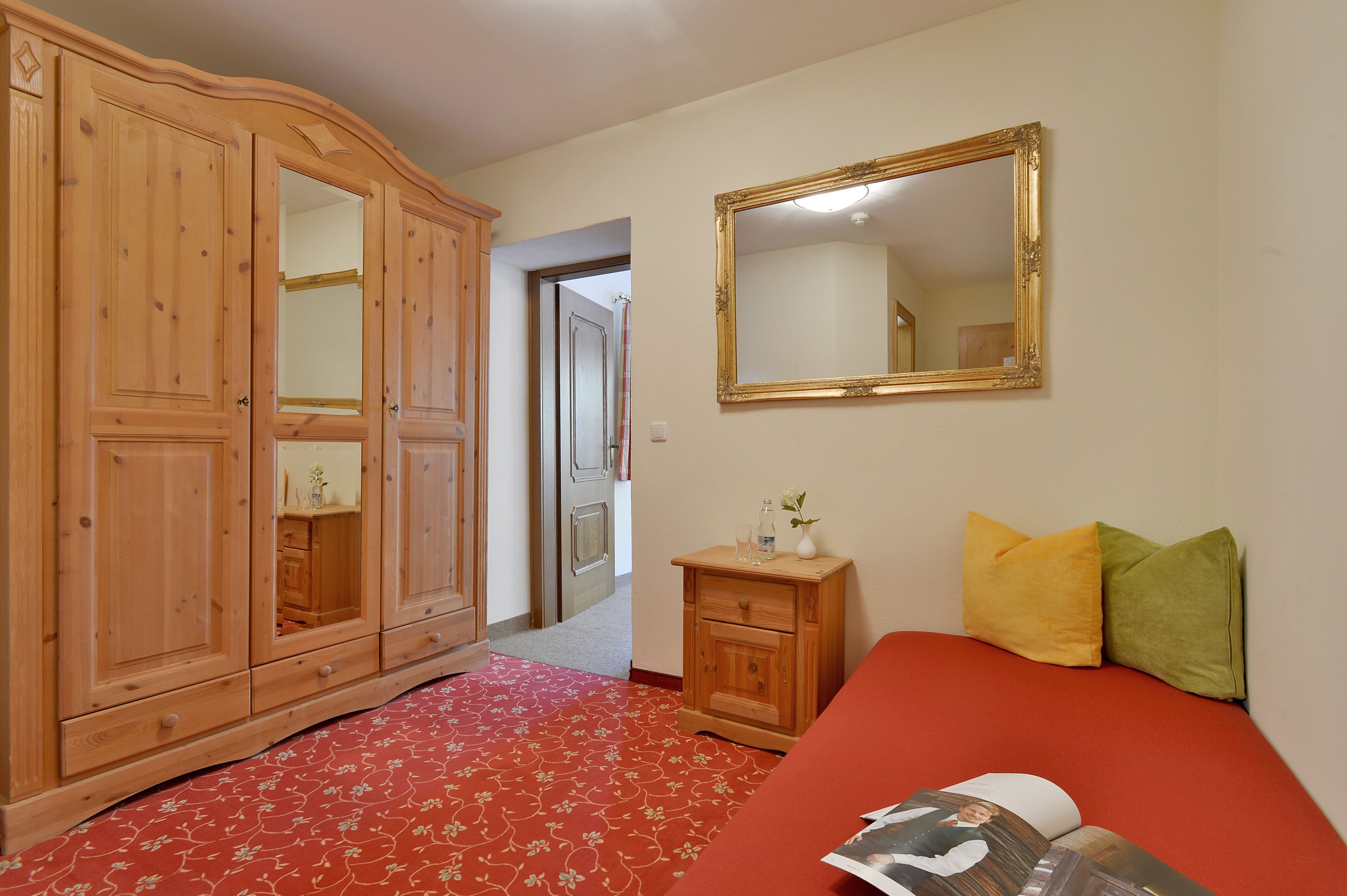 Une chambre avec une armoire en bois, un miroir, une table de chevet ornée d’un vase et de verres, et une couvre-lit rouge.