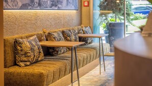 Uma área de estar aconchegante com um sofá estampado e duas mesinhas, um quadro na parede e um vaso de planta lá fora.
