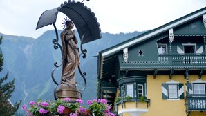 Uma estátua com guarda-chuva e flores, com uma montanha e um edifício com varandas ao fundo.
