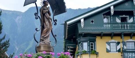 A statue with an umbrella and flowers, set against a mountain backdrop and a building with balconies.