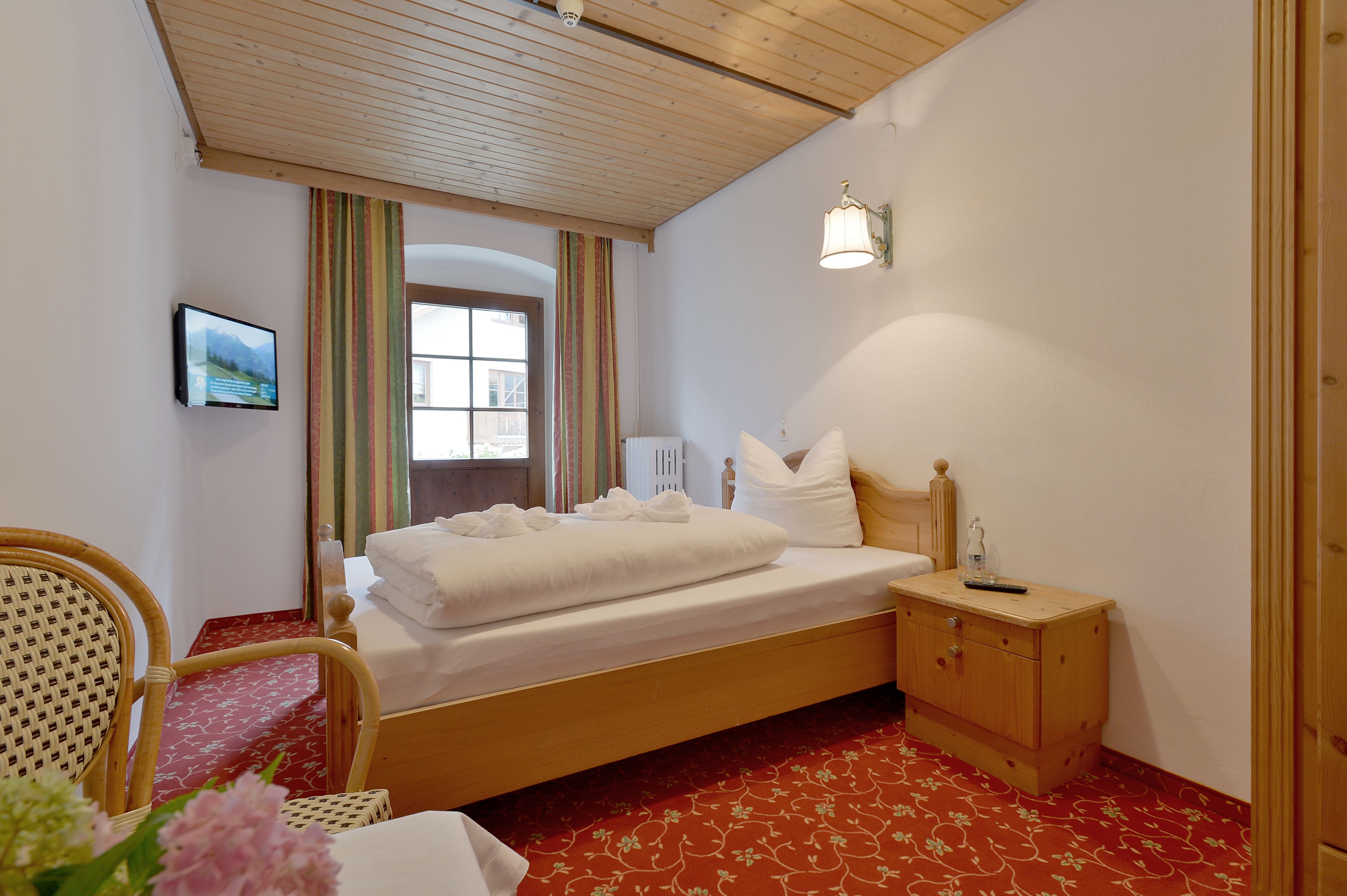 A neatly made bed with white linens, a wooden nightstand with a lamp, a wicker chair, and a small table with a vase of flowers.