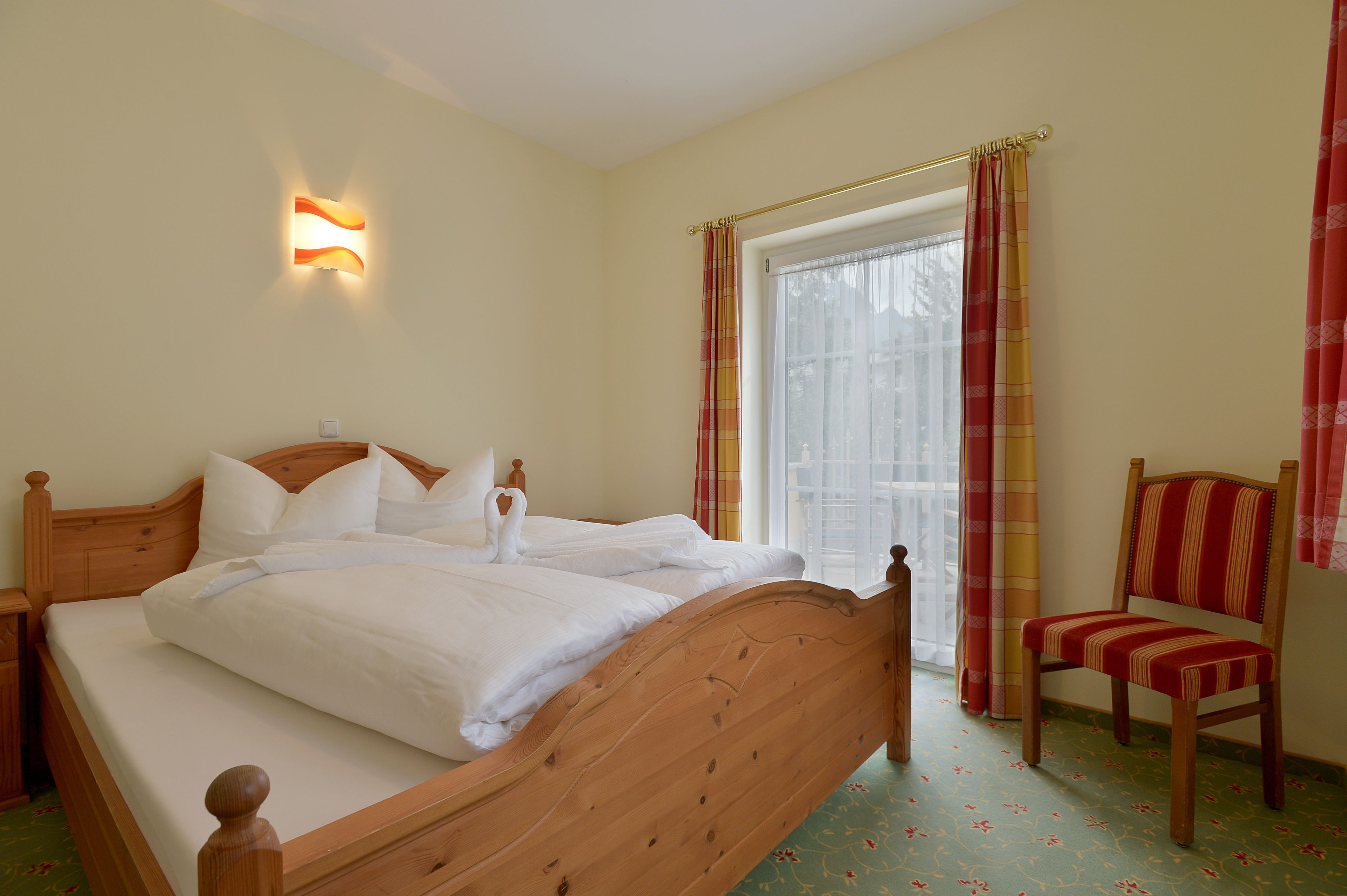 A double bed with white linens, a wooden headboard, and a nightstand. There is a window with curtains, a chair, and a wall-mounted light fixture.