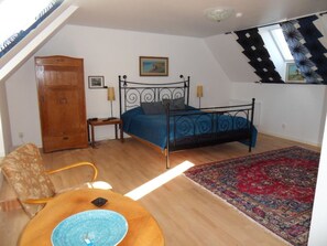 A bedroom with a bed, a wooden wardrobe, a chair, a table with a blue bowl, and a patterned rug.