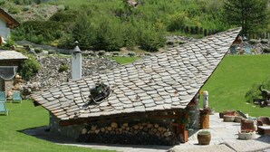 A small stone-roofed cabin with a log storage area, surrounded by a well-maintained garden and a stone wall. - Notre Maison Eco Wellness Hotel (Cogne)