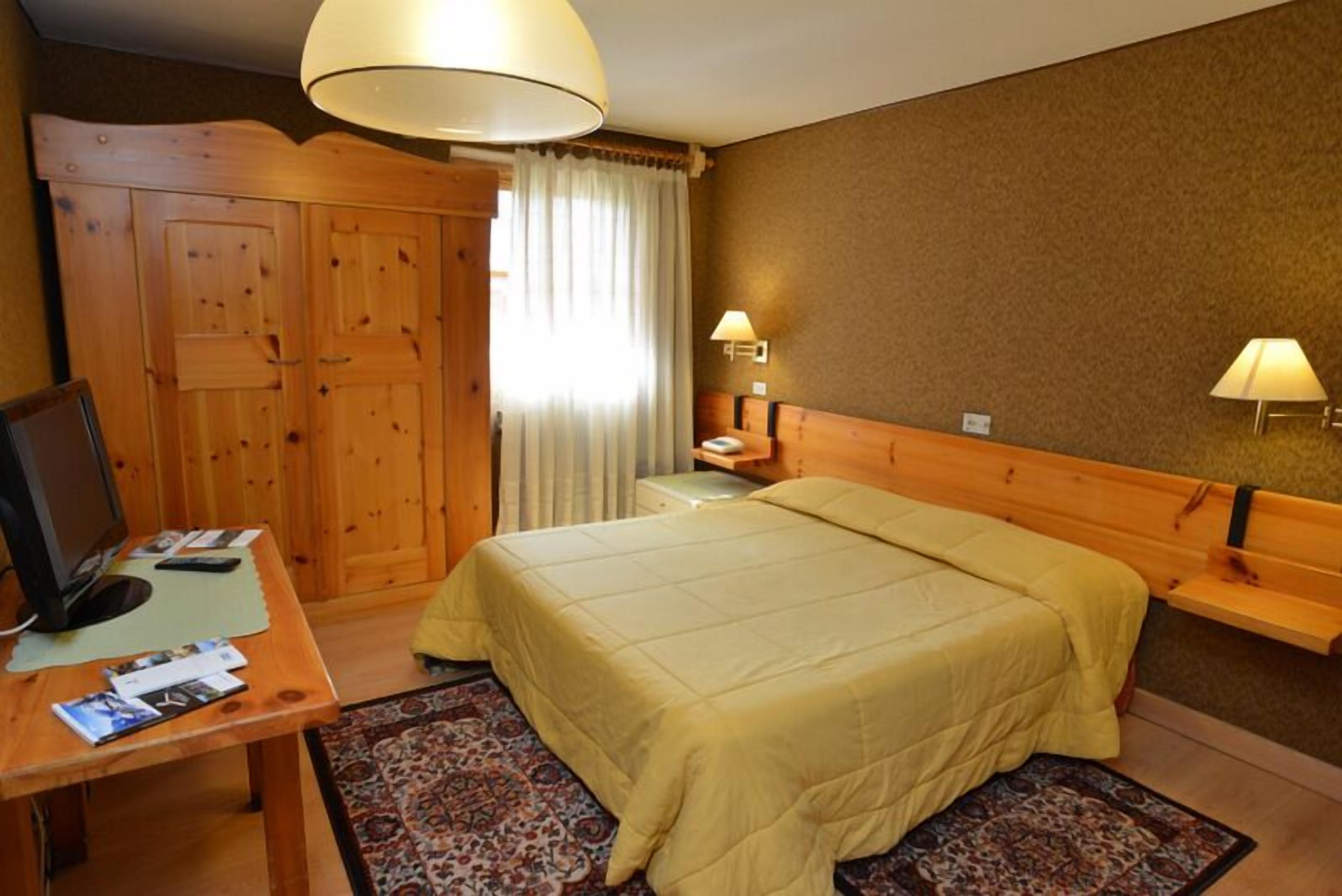 Una camera d'albergo con un letto in legno, una scrivania con un computer, una lampada e un armadio.
