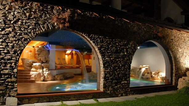 An outdoor spa area with a pool, stone arches, and a waterfall. - Notre Maison Eco Wellness Hotel (Cogne)