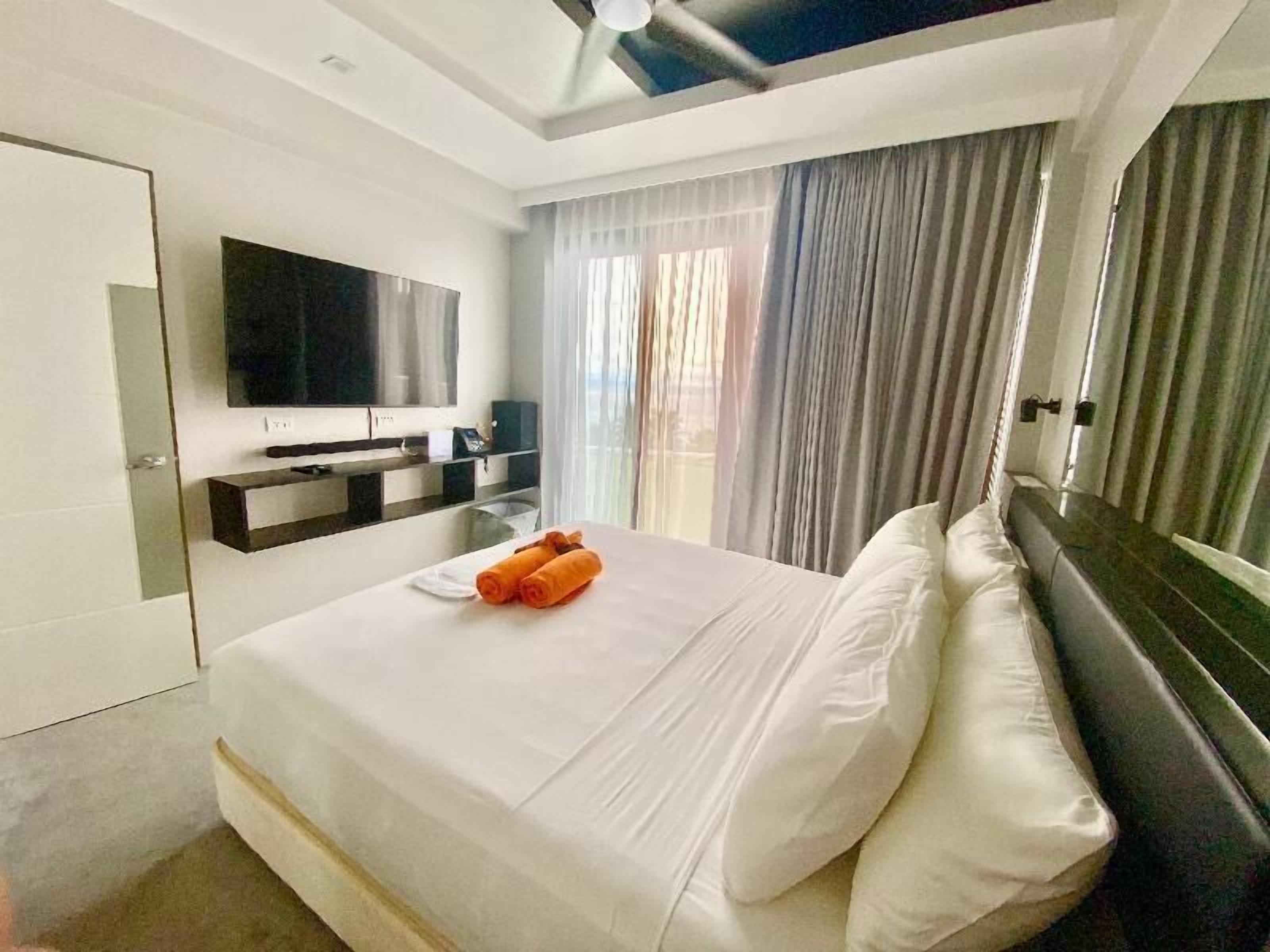 Quarto de hotel moderno com uma cama grande, uma televisão de tela plana na parede, uma mesinha de cabeceira e uma janela com cortinas.