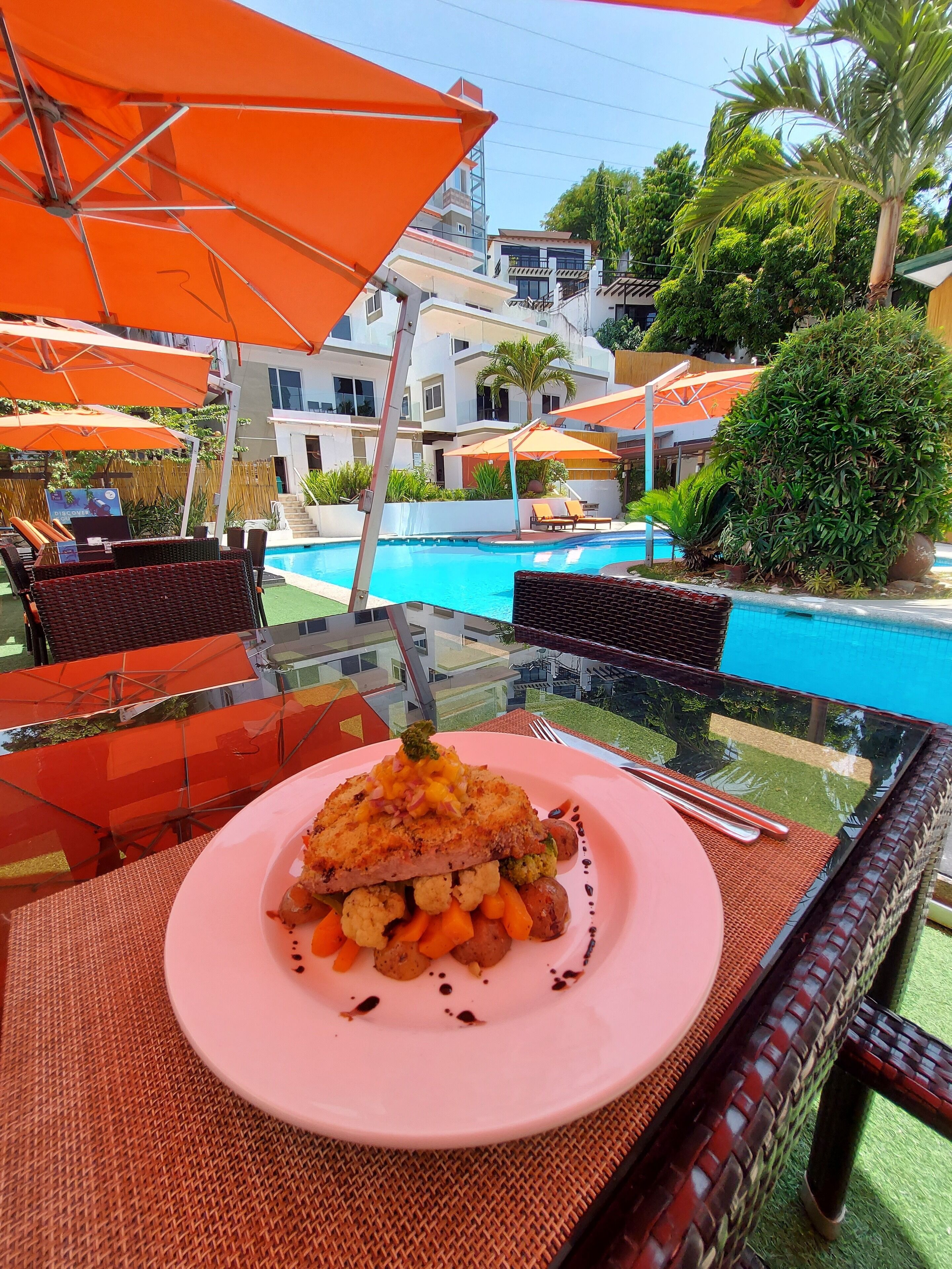 A dining table with a plate of food, orange umbrellas, and a pool in the background.