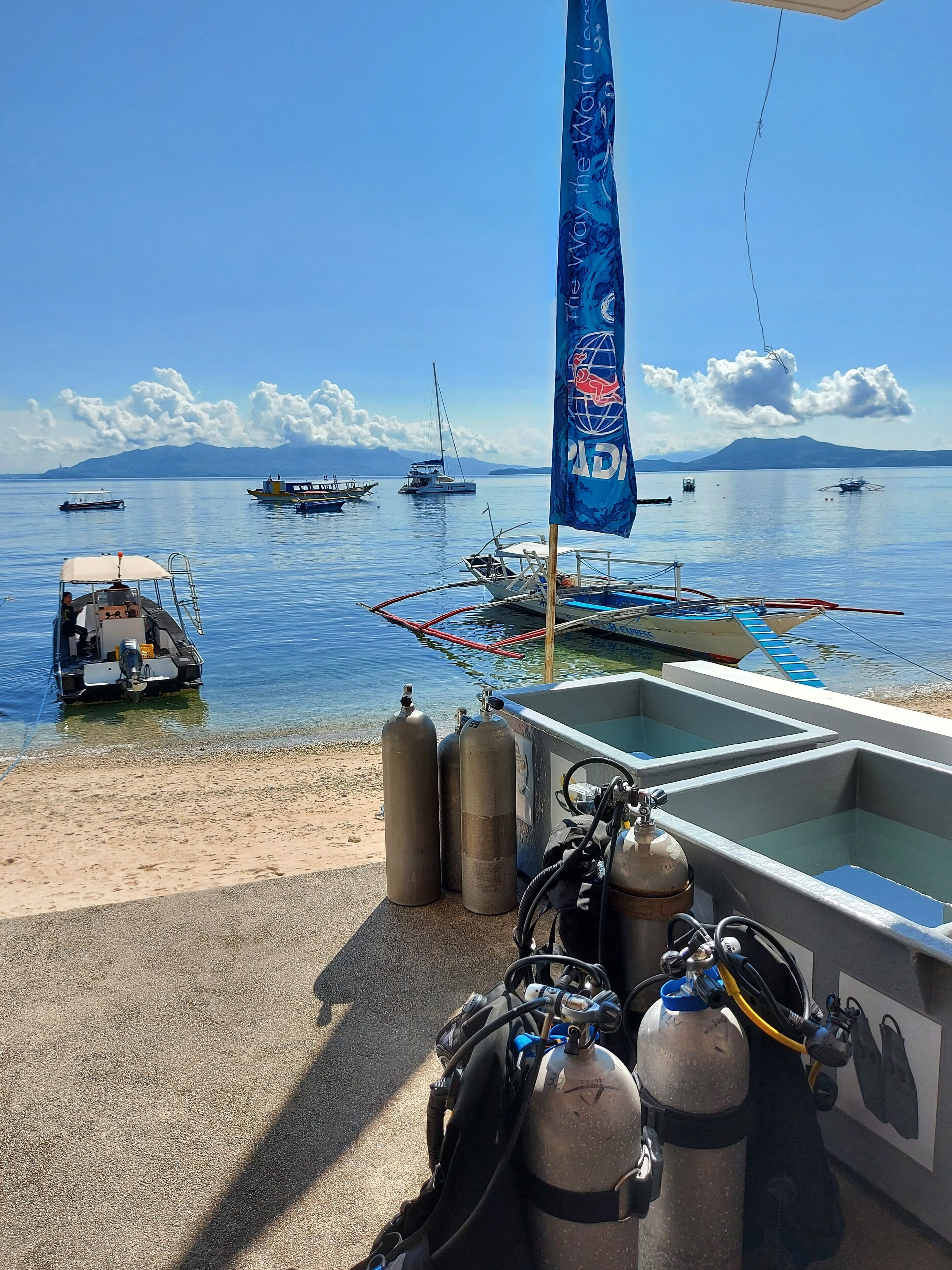 A boat docked at a pier with a flag, scuba tanks, and a clear view of the sea with boats and mountains in the distance.