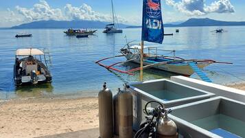 A boat docked at a pier with a flag, scuba tanks, and a clear view of the sea with boats and mountains in the distance.