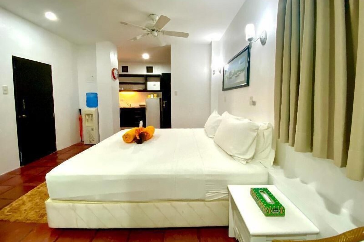 A hotel room with a bed, a small bedside table, a ceiling fan, a kitchen area in the background, and a door.