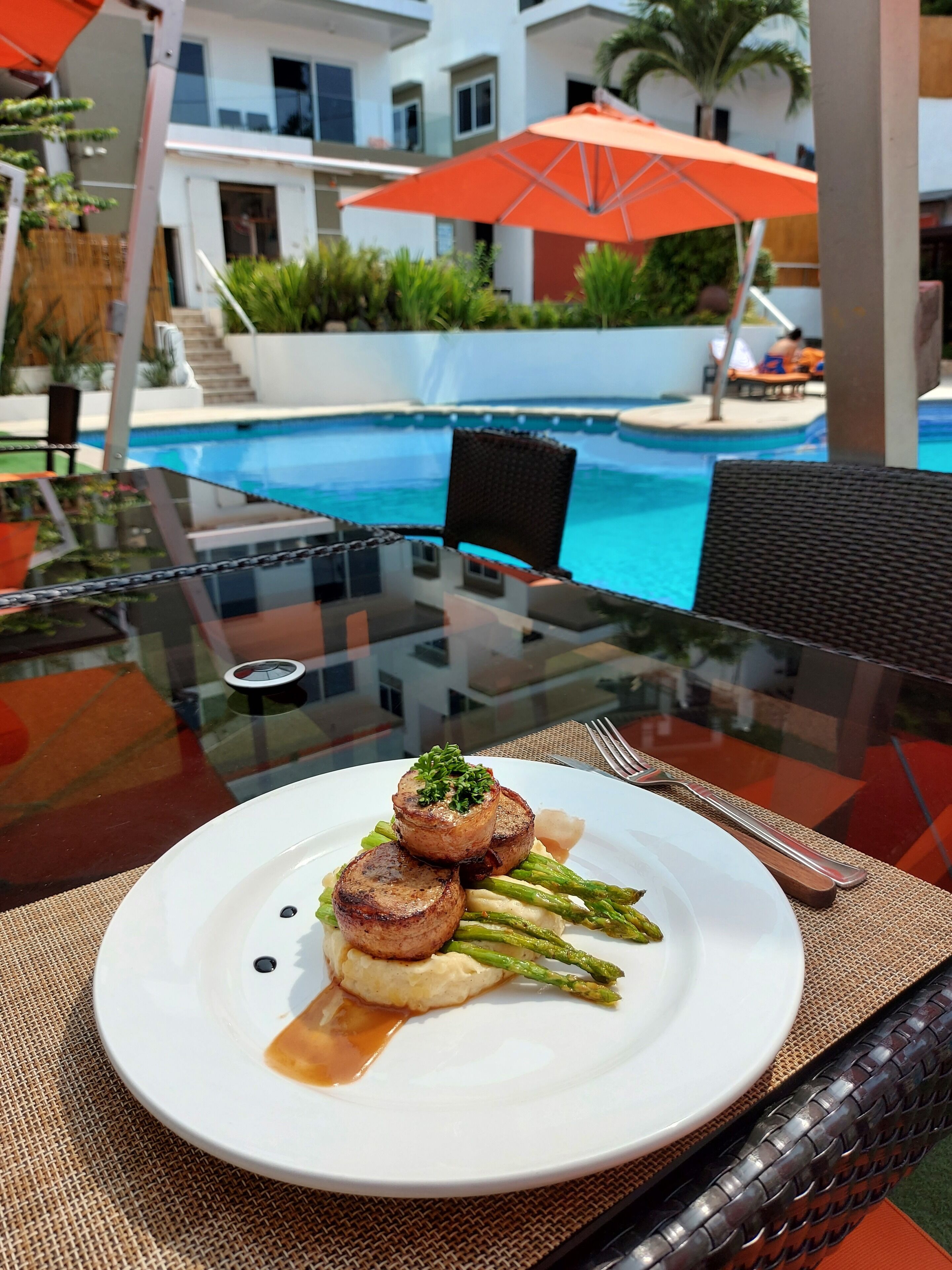 A dining table with a plate of food, a glass table, and a pool in the background.