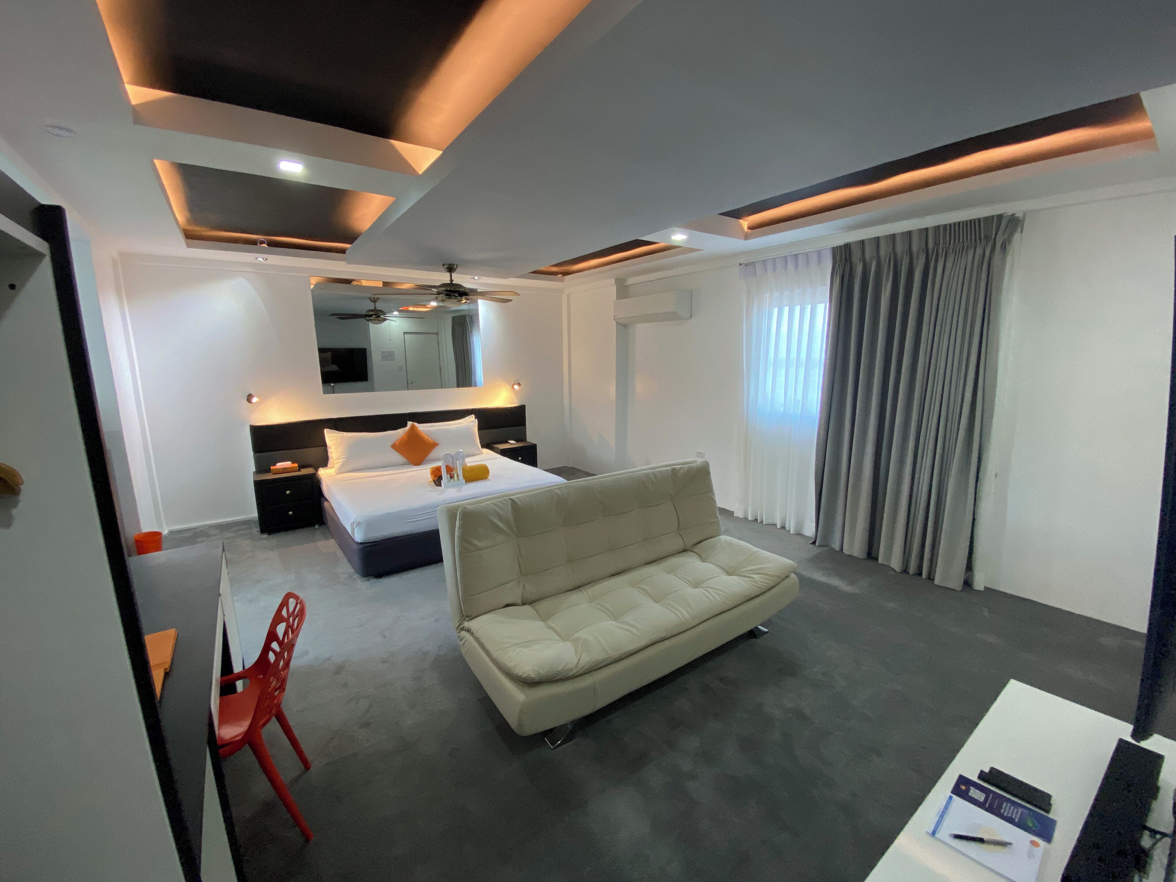 A modern hotel room with a bed, a sofa, a red chair, a desk, and a large window with curtains.