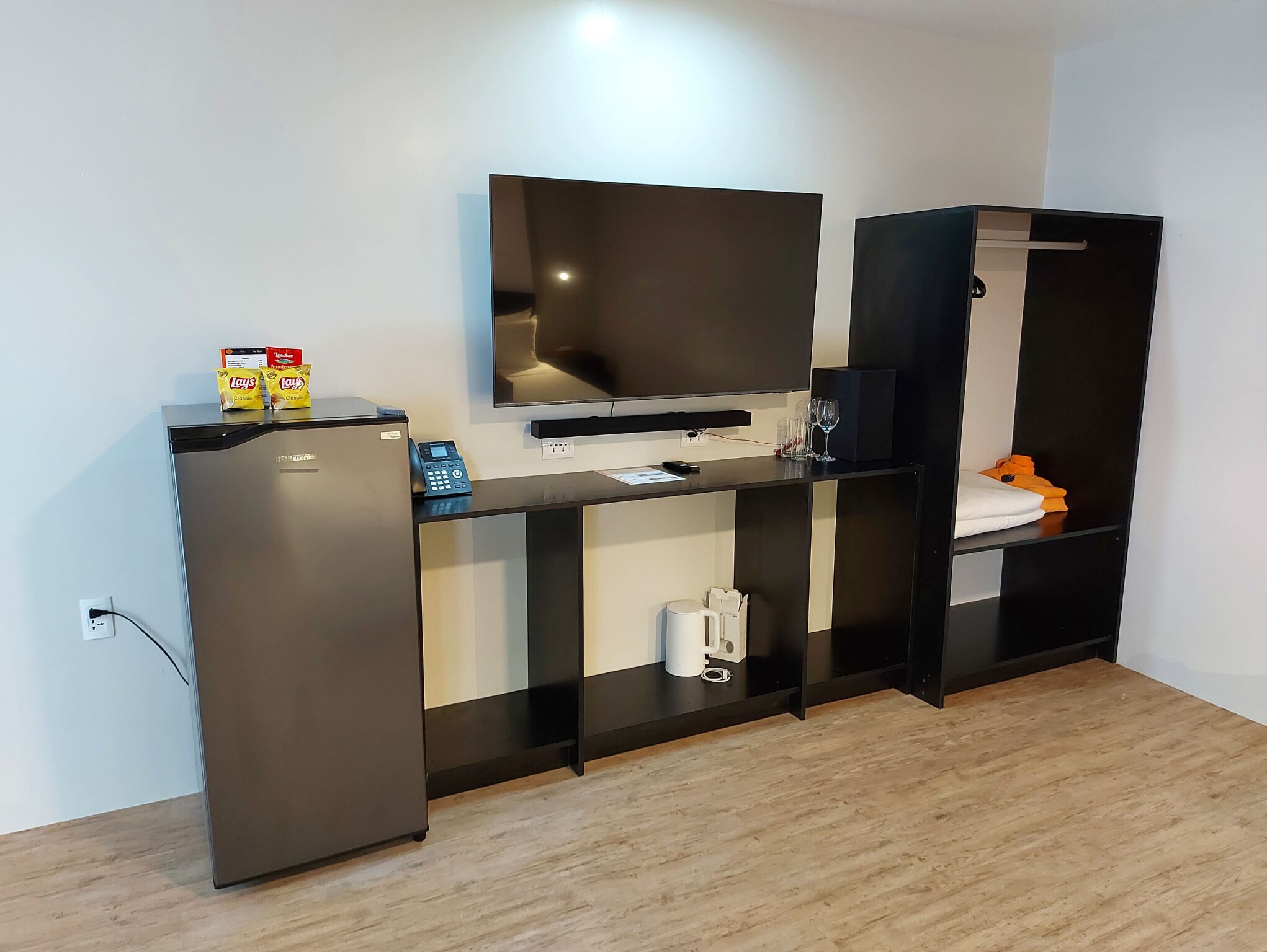 A compact living space with a refrigerator, a flat-screen TV, a small kitchenette, and a wardrobe.