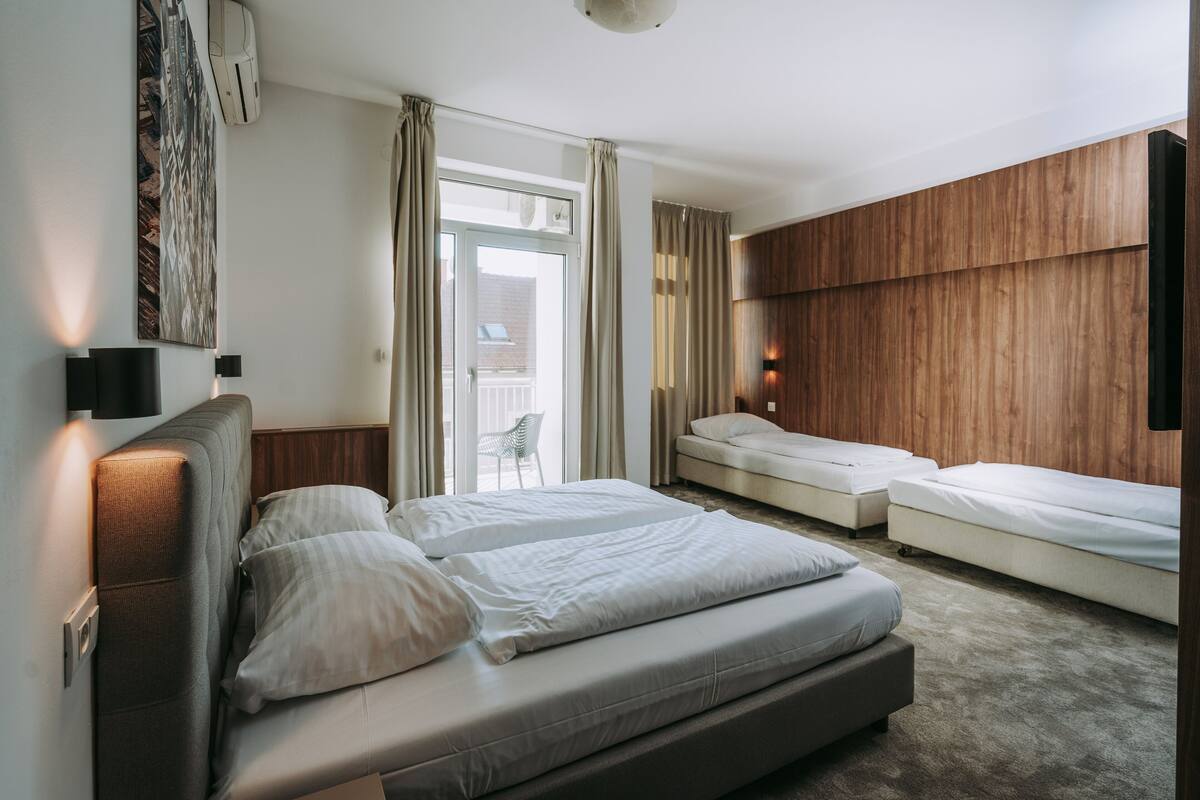 A hotel room with a large bed, a smaller bed, a sofa bed, a wooden panel wall, a ceiling light, and a window with curtains.